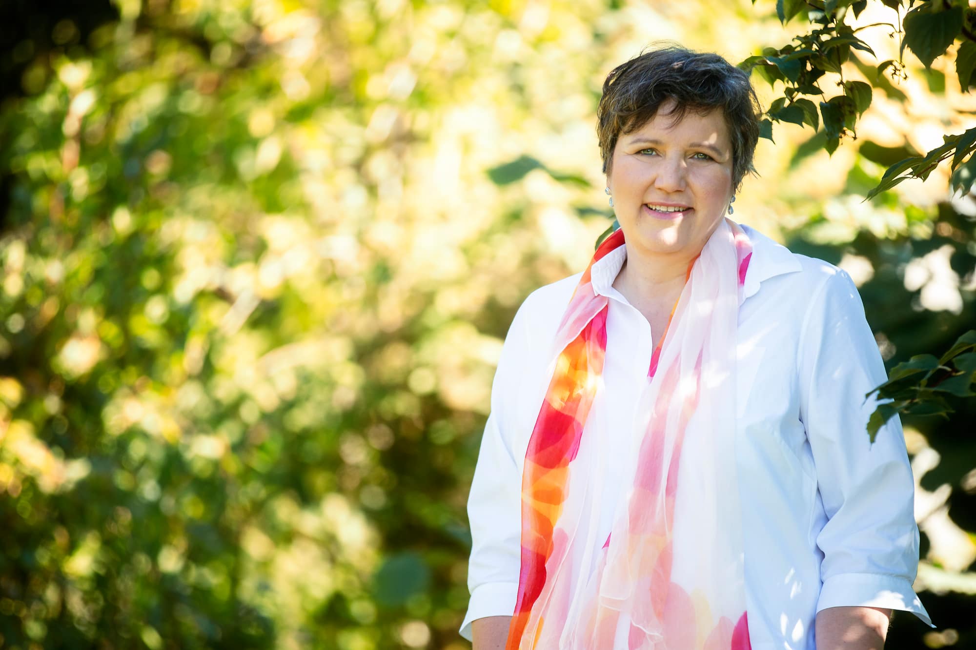 Gabriele Kirchmann lächelt in weißer Bluse mit buntem Seidenschal vor sonnigem Blattgrün